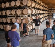 Eight people of varying ages and races gather around a tour guide, who is talking about the distillery process on a tour of J. Henry Bourbon Distillery. They are all standing in front of large racks of hundreds of barrels.