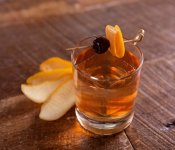 A cocktail in a whiskey glass with a fruit peel and cherry garnish on top, fastened to the glass with a skewer. The cocktail is sitting on a wooden table.