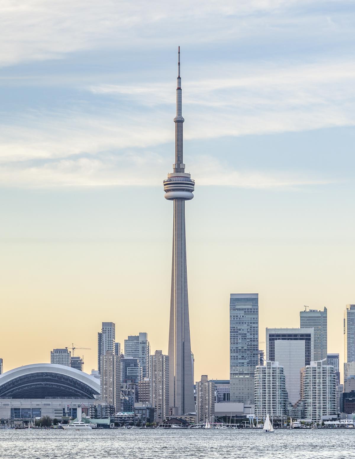 The CN Tower, a tall, iconic red and white tower, dominates the Toronto skyline. The tower stands tall above the city's skyscrapers, with Lake Ontario in the foreground and a dramatic  sunset sky in the background.