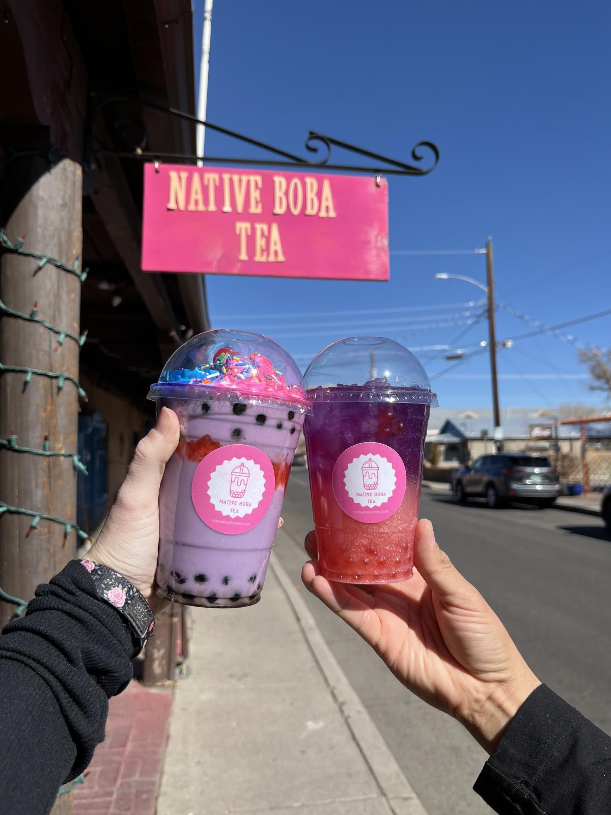 Two people "cheers" with two drinks in front of the Native Boba Tea sign.