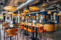 The Astronomer bar area at the Hyatt Centre Cambridge. The bar is in the background with dark blue walls & a wrap around light wood colour panel. There are 2 high tables with orange bar stools on a tiled hard floor