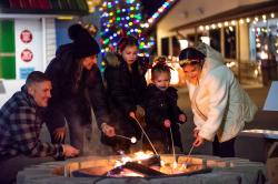 Picture of group of people huddled around a fire pit roasting marshmallows at the Nickle Plate Christmas Village s'more station!