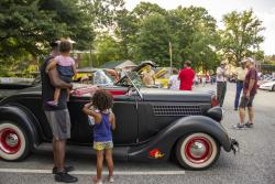 A man holding a young toddler and his preschool age child look at an antique car while other people mill around at a car show.