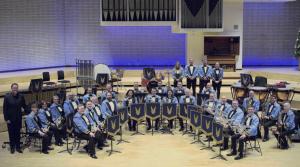 The KNDS Fairey Band sit in rows with their instruments and sheet music on stands, in a room which looks like a recording studio