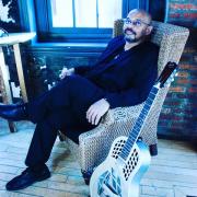 Norman Taylor poses in a wicker chair next to a guitar.