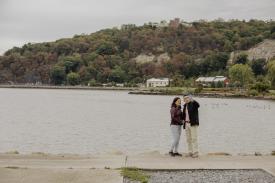 A couple stands taking a selfie in front of the Hudson River at Riverfront Green Park. Both the sky and water behind them look grey.