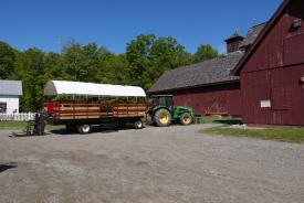 A red and brown covered wagon is attached to a green tractor. There is a ramp at the back of the wagon.