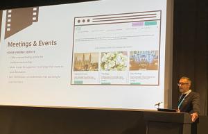 Black background with a white projection screen in foreground. On the screen is the home page of Meet Cambridge with a white man to the side behind a lectern