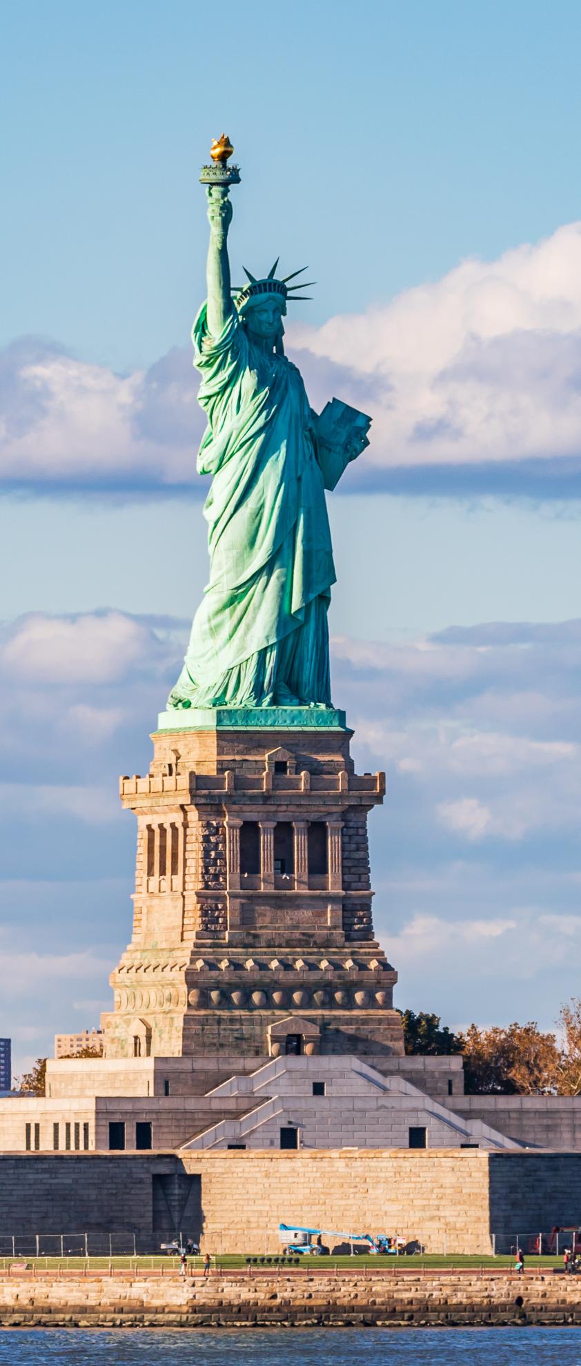 The Statue of Liberty, a colossal copper statue of a woman holding a torch and a book, stands on Liberty Island in New York City. The statue is surrounded by water and skyscrapers in the  background.