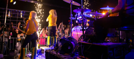View from behind the stage showing a singer, guitarist, drum set, and audience during a performance.