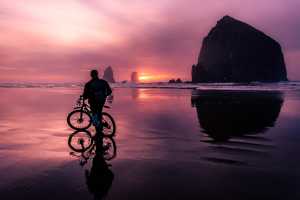 A person with a bicycle on the beach at sunset. Bicycle, person, and Haystack Rock all in silhouette.