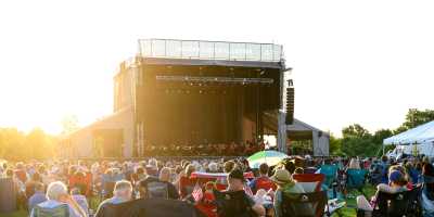 Crowd seated on lawn chairs watching the Louisville Orchestra perform during the Play America concert event at the Jasteka Cultural Center outdoor stage at sunset.
