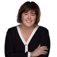 Lori Stinson is a Caucasian female with chin-length brown hair and blue eyes, she is smiling at the camera with arms crossed wearing a black and white button up sweater