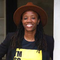 Nanyamka Moore is a black female, she is smiling at the camera wearing a brown felt hat and black and yellow sweatshirt