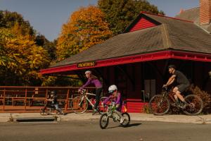 A family consisting of a mom, dad, young daughter and son, bike ride with helmets on past an older depot train station building.