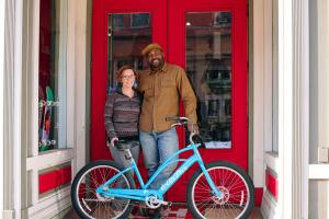 A man and woman stand in front of a red door holding a bright blue bike.