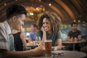 A black woman who has a glass of wine smiles at an Asian man sitting with her who drinks a beer.