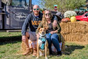 A man and woman are shown sitting on a hay bale posing for a photo with their brown and white dog. An antique truck with a Dogfish Head Brewery logo printed on it sits in the background.