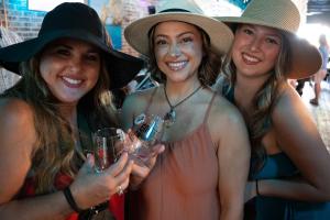 Three women wearing sun hats and smiling show their wine glasses from the event day