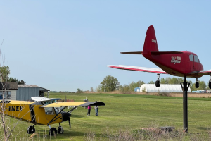 A yellow airplane sits on a grassy airfield beside a red aircraft displayed on a raised stand under a clear blue sky.