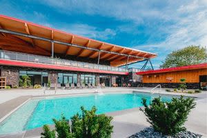 An outdoor swimming pool beside a modern building with large windows, a wood-accented roof, lounge chairs, and landscaped greenery.