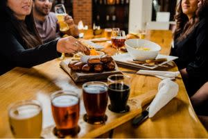 Group of people sharing appetizers and a flight of craft beers at a wooden table, dipping pretzels into sauce in a cozy restaurant setting.