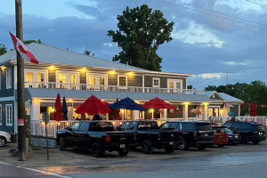 A two-storey clubhouse with a brightly lit outdoor patio, colourful umbrellas, a Canadian flag, and vehicles parked in front at dusk.