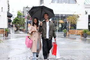 A smiling couple walking through the McArthurGlen West Midlands shopping village, Staffordshire, with plenty of shopping bags, on a rainy day