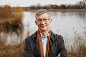 Press shot of Nick Higham, standing in front of a lake and smiling at the camera