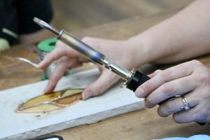 Picture of hands making glass art. One hand placing glass and the other hand holding a soldering iron at a STEAM Innovation Lads Art Glass Class.