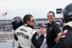 The Governor of Pennsylvania, Josh Shapiro, looking back at a camera, smiling while talking with a race car driver with ear plugs in and holding a helmet