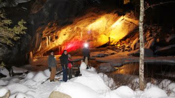 A group of four people stand on snow near a large, illuminated cave entrance at night. The scene conveys adventure and exploration in a winter landscape.