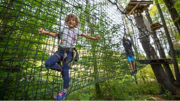 Young girl with brown hair, wearing a harness, confidently crossing a rope net bridge in a forest adventure park, with trees surrounding her.