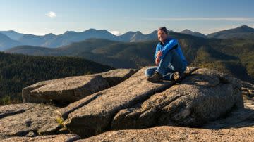 A person in a blue jacket sits on a rocky ledge, surrounded by a vast mountainous landscape under a clear blue sky, evoking tranquility and adventure.