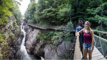 A couple stands on a wooden bridge overlooking a waterfall with lush greenery and rocky cliffs. They appear happy and the scene feels serene.