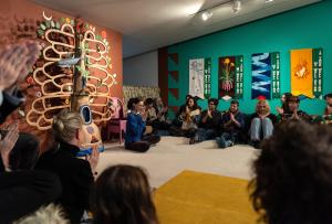 A crowd seated in a circle hold their hands in a prayer pose as colorful textile banners hang above them and an abstract tree-like sculpture rests next to them.