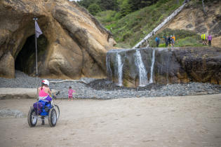 A person rides their trike on the beach and stops to watch the waterfall at Hug Point State Park.