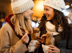 Two women smile while eating food at an outdoor holiday market