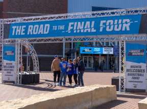 Four people pose under a large sign that reads "Road to the Final Four" outside of INTRUST Bank Arena during the NCAA Men's Division 1 Basketball Championship.
