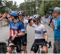 A group of 3 female cyclists smile and high five each other as a man makes an announcement with a microphone.