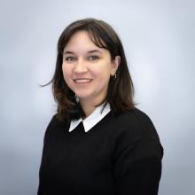 A professional headshot of a woman with shoulder-length dark hair, wearing a black sweater over a white collared shirt. She has a friendly smile and is posed against a light blue background