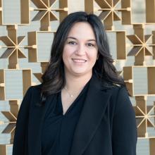 A smiling woman with shoulder-length dark hair stands in front of a decorative geometric metal wall, wearing a black blazer and blouse with a delicate necklace, softly lit by natural light.