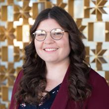 A smiling woman with long dark wavy hair and round glasses stands in front of a decorative geometric metal wall, wearing a burgundy blazer over a patterned top, softly lit by natural light.