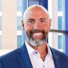 A smiling man with a bald head and gray beard wears a blue suit and white shirt, standing in front of tall windows with a blurred view of a modern building behind him.