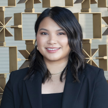 A smiling woman with shoulder-length dark hair stands in front of a decorative geometric metal wall, wearing a black blazer and top with subtle jewelry, softly lit by natural light.