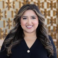 A woman with long dark hair and soft highlights smiles warmly while wearing a black top, standing in front of a softly blurred geometric metal wall in warm tones.