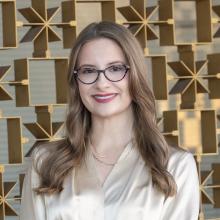 A woman with long light brown hair and glasses smiles while wearing a cream blouse and layered gold necklaces, standing in front of a warm-toned geometric metal wall.