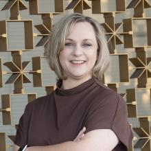 A smiling woman with short blonde hair stands with her arms crossed in front of a decorative geometric metal wall, wearing a brown top and a smartwatch, softly lit by natural light.