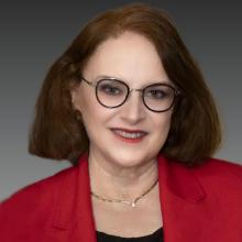 Beth Duke headshot - caucasian woman with short red hair wearing a bright red coat with black round glasses