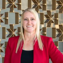 A woman in a red blazer and black top smiles confidently with one hand on her hip, standing in front of a geometric metal wall with warm tones.